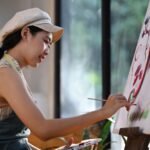 Happy young asian woman painting picture on canvas with oil paints in her studio.