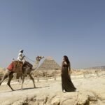 A man riding a camel and a woman having a picture to the great pyramid of Giza as background as one of the things to do in Egypt.