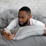 Confused black man looking at phone in bed