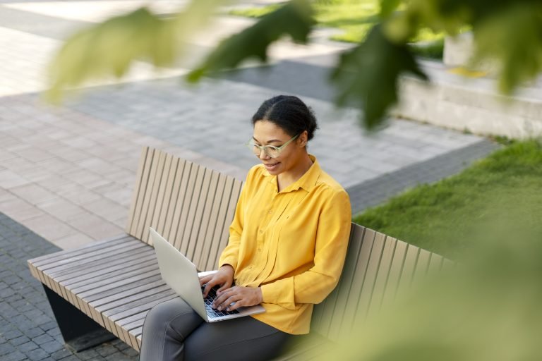 Smiling African American businesswoman using laptop computer working online, sitting on bench