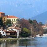 One of the best places in Northern Italy is a picturesque mountain town of bassano del grappa with clustered houses on a hillside overlooking a river valley, with mountains in the background under a blue sky.