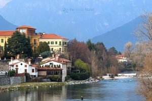 One of the best places in Northern Italy is a picturesque mountain town of bassano del grappa with clustered houses on a hillside overlooking a river valley, with mountains in the background under a blue sky.
