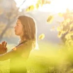 A woman in a black shirt with her hands together meditating in a nture retreat.