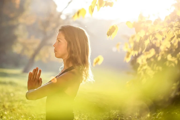 A woman in a black shirt with her hands together meditating in a nture retreat.