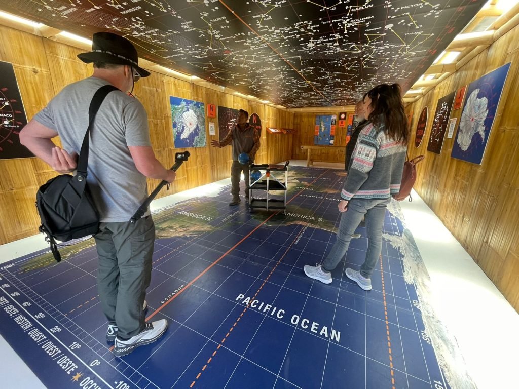 Visitors inside a museum room with a world map on the floor, star constellations on the ceiling, and educational displays on the walls.
