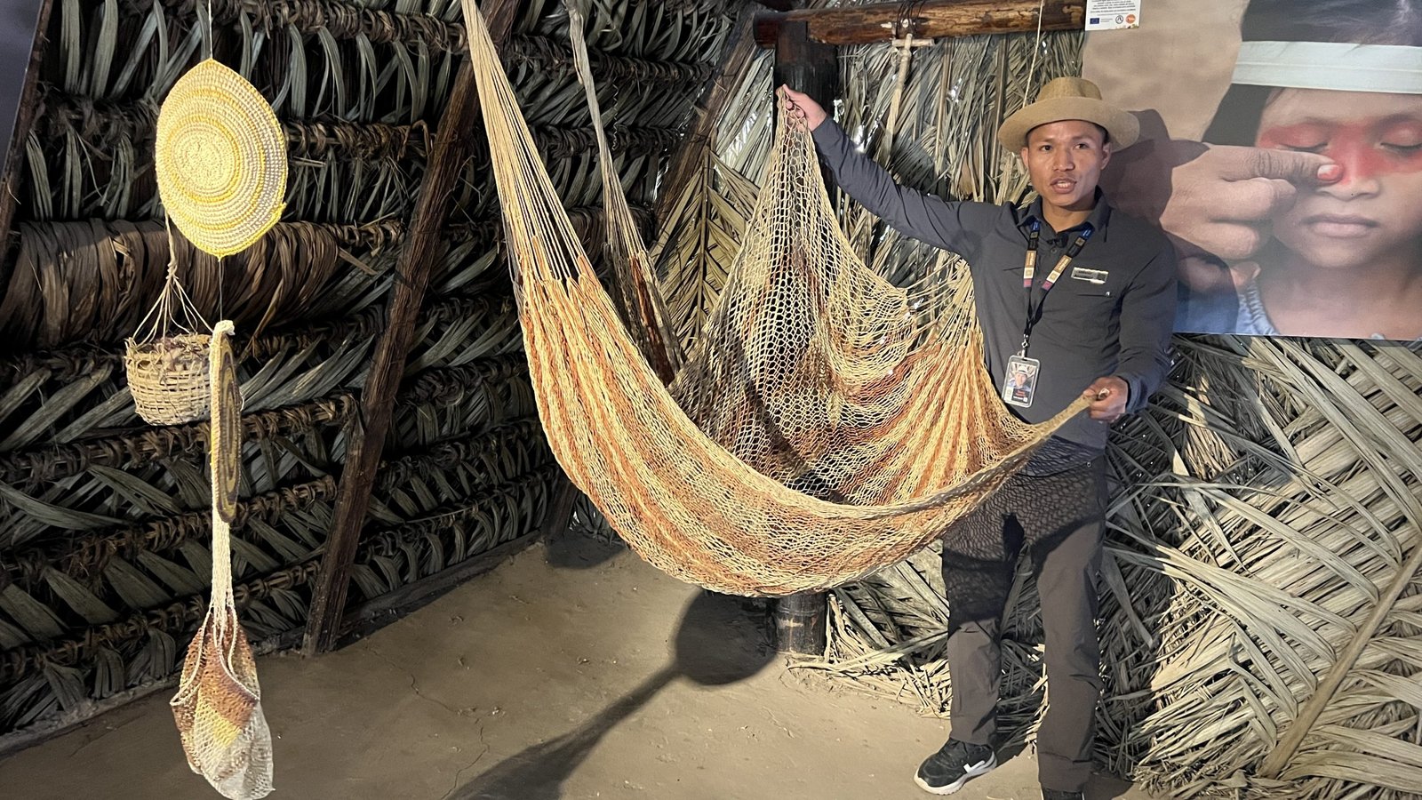 Tour guide presenting a woven hammock in a traditional hut made of palm leaves, with cultural items and a photo backdrop.