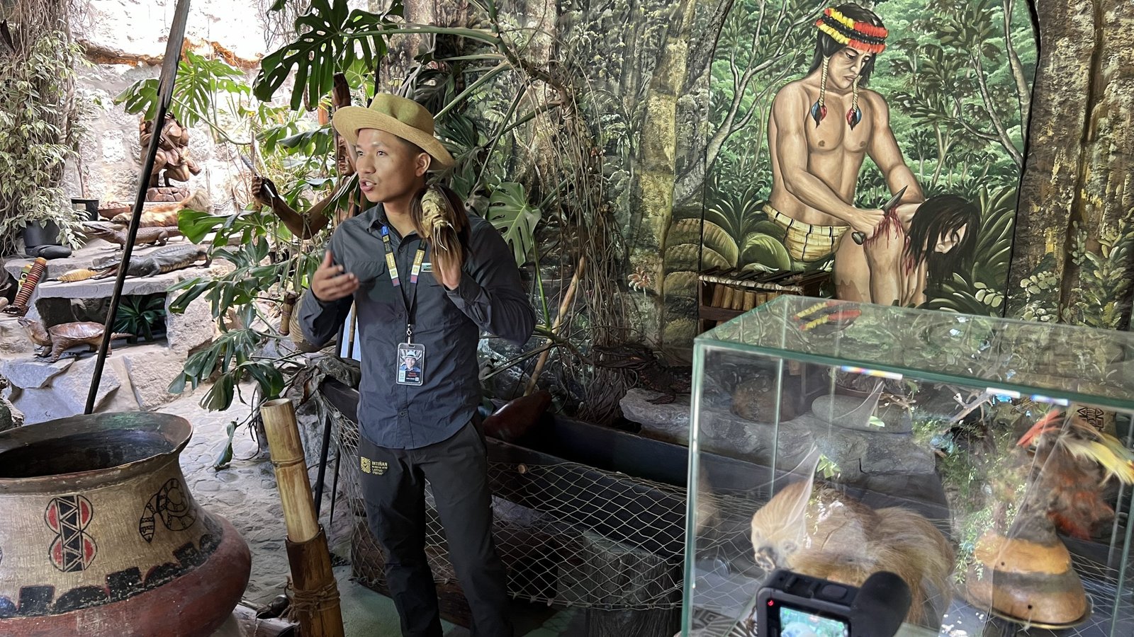 Tour guide wearing a straw hat, speaking in a cultural exhibit with jungle-themed murals, traditional artifacts, and a display case featuring indigenous items.