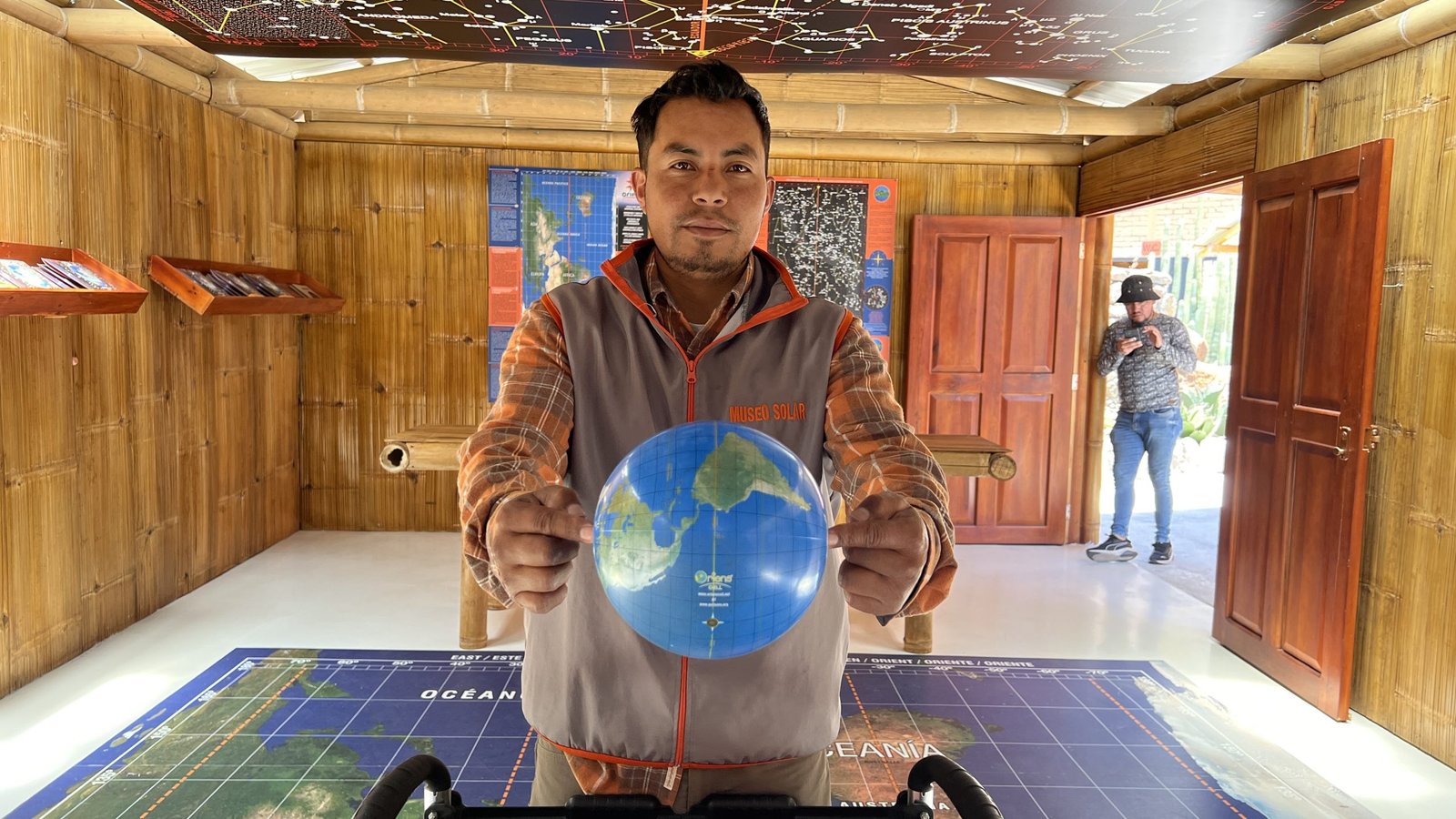 Museum guide holding a small globe, standing in a bamboo-paneled room with maps and educational exhibits.