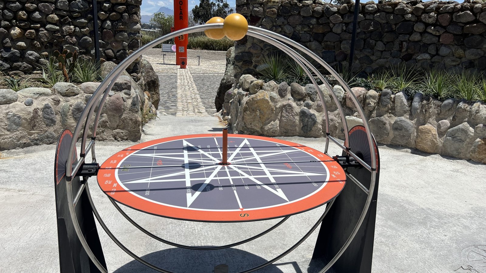 Sundial with a circular compass and metal framework, set in an outdoor exhibit with stone walls in the background.