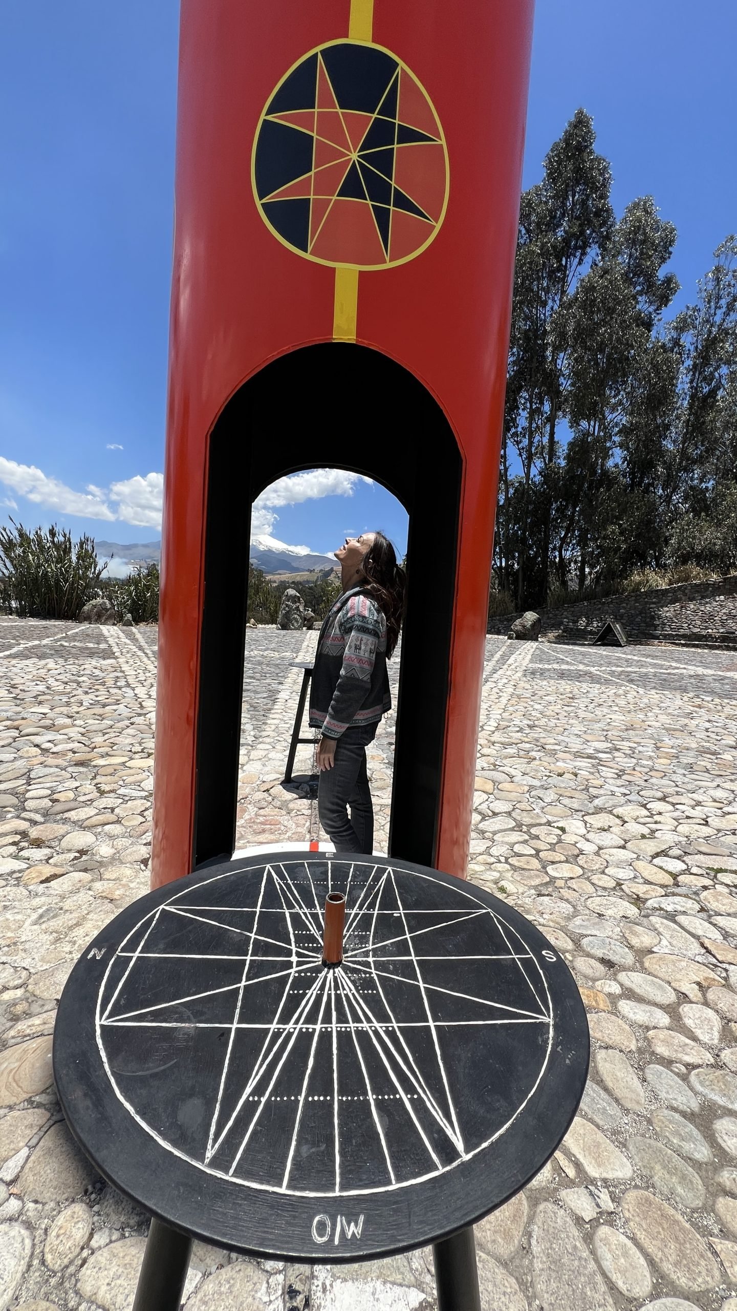 Woman standing inside a red equatorial marker, with a sundial in the foreground and trees in the background.