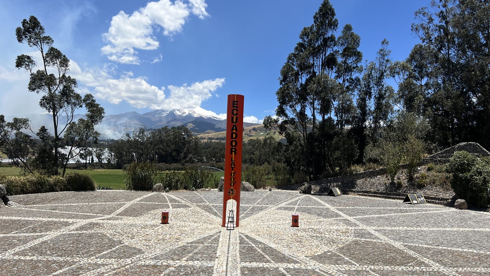 Stone plaza with an orange "Ecuador Latitude 0°0'0''" marker surrounded by trees, mountains, and a clear blue sky.
