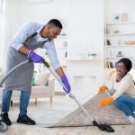 Positive black couple cleaning up together, vacuuming under carpet at their house, copy space