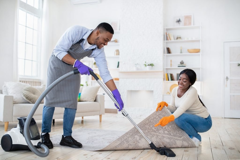 Positive black couple cleaning up together, vacuuming under carpet at their house, copy space