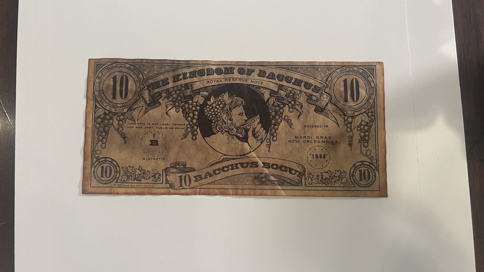 A vintage 1968 Bacchus Mardi Gras paper throw resembling a ten-dollar bill, featuring "The Kingdom of Bacchus" and an illustration of Bacchus surrounded by grapevines. The note is aged and worn, placed on a white surface. The caption mentions that these paper throws were short-lived due to difficulty in tossing.