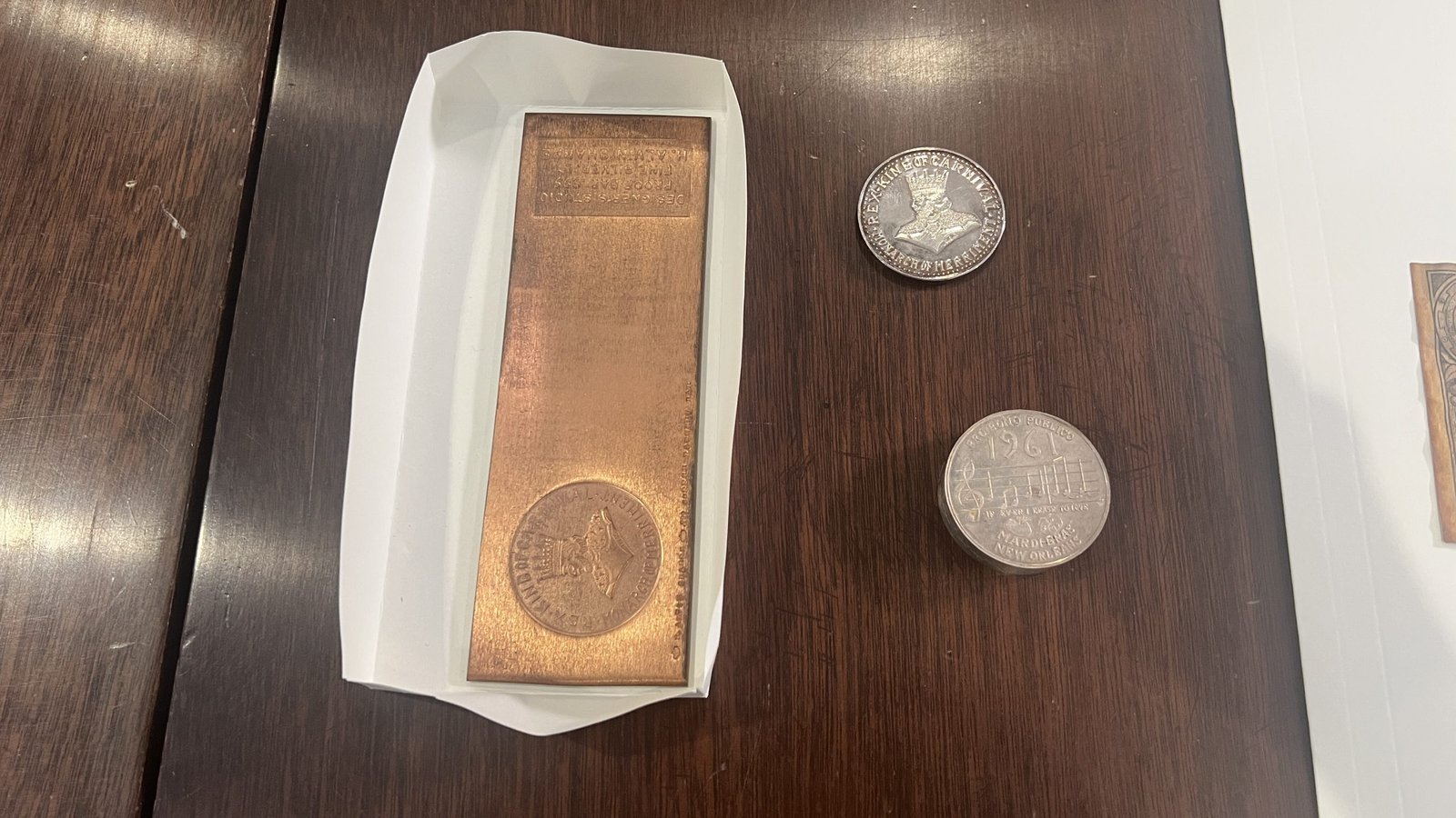 A rectangular copper-toned Mardi Gras doubloon in a white paper holder, placed on a wooden surface. To the right, two round doubloons—one silver and one bronze—feature embossed designs. The quote above describes the anodized tin material and its lightweight nature, making it easy to throw during Mardi Gras celebrations.