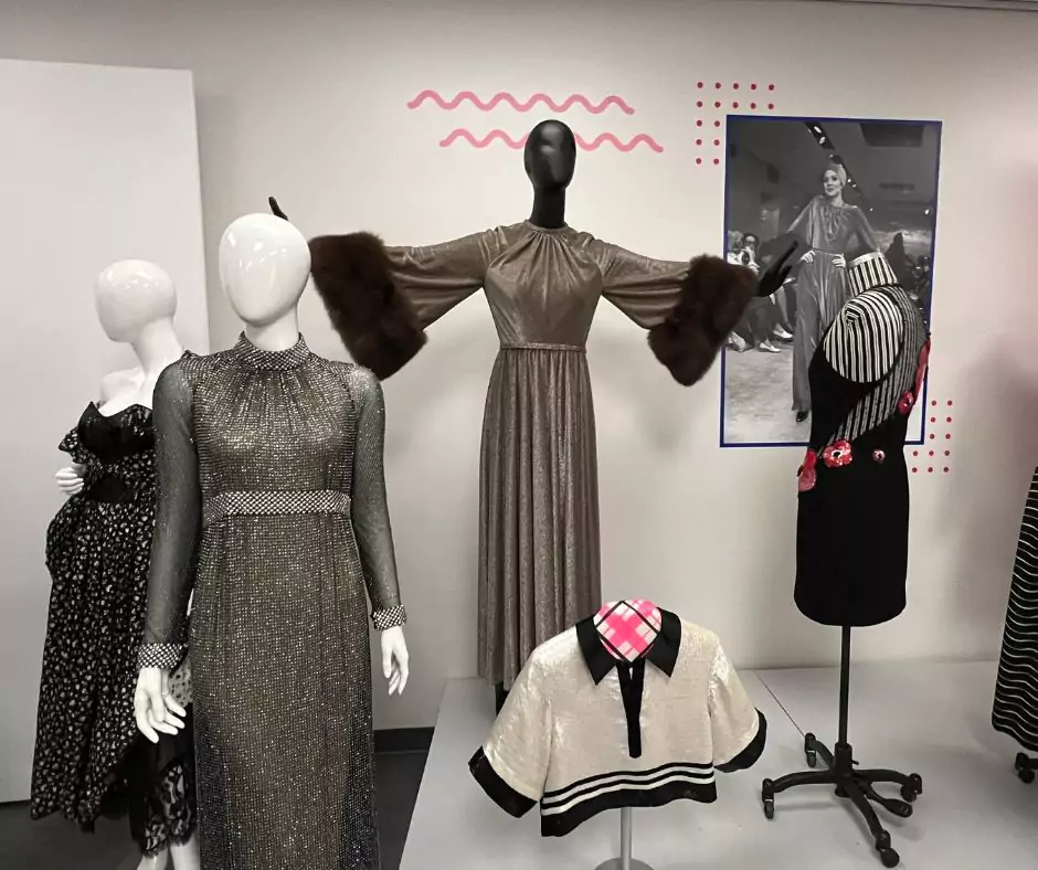 A Geoffrey Beene exhibit featuring elegant eveningwear, including a metallic gown with dramatic fur-trimmed sleeves, a high-neck shimmer dress, and a black dress with floral embroidery. A framed runway image adds historical context.