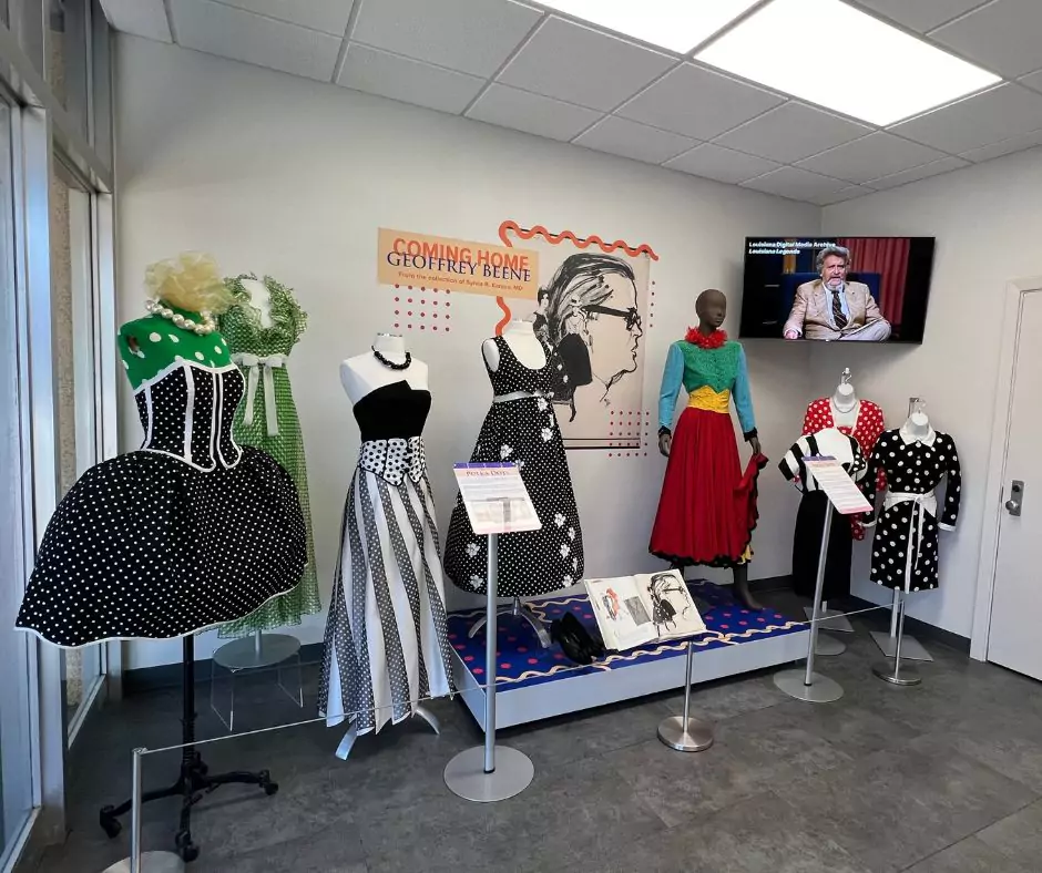 A collection of Geoffrey Beene dresses on mannequins, showcasing polka-dot patterns, bold color blocking, and vintage silhouettes. A video screen in the background plays an interview about Beene’s contributions to fashion.