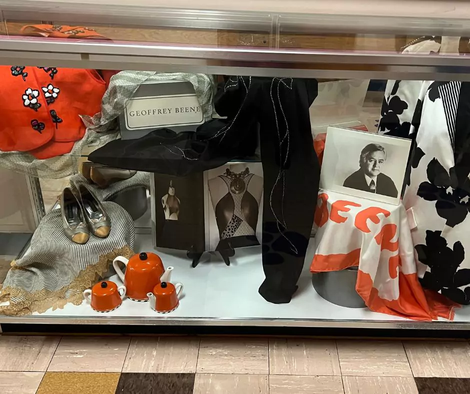 A glass display case showcasing Geoffrey Beene memorabilia, including a designer vest, silver shoes, a framed portrait of Beene, and red ceramic teapots. The exhibit highlights his influence on both fashion and design.