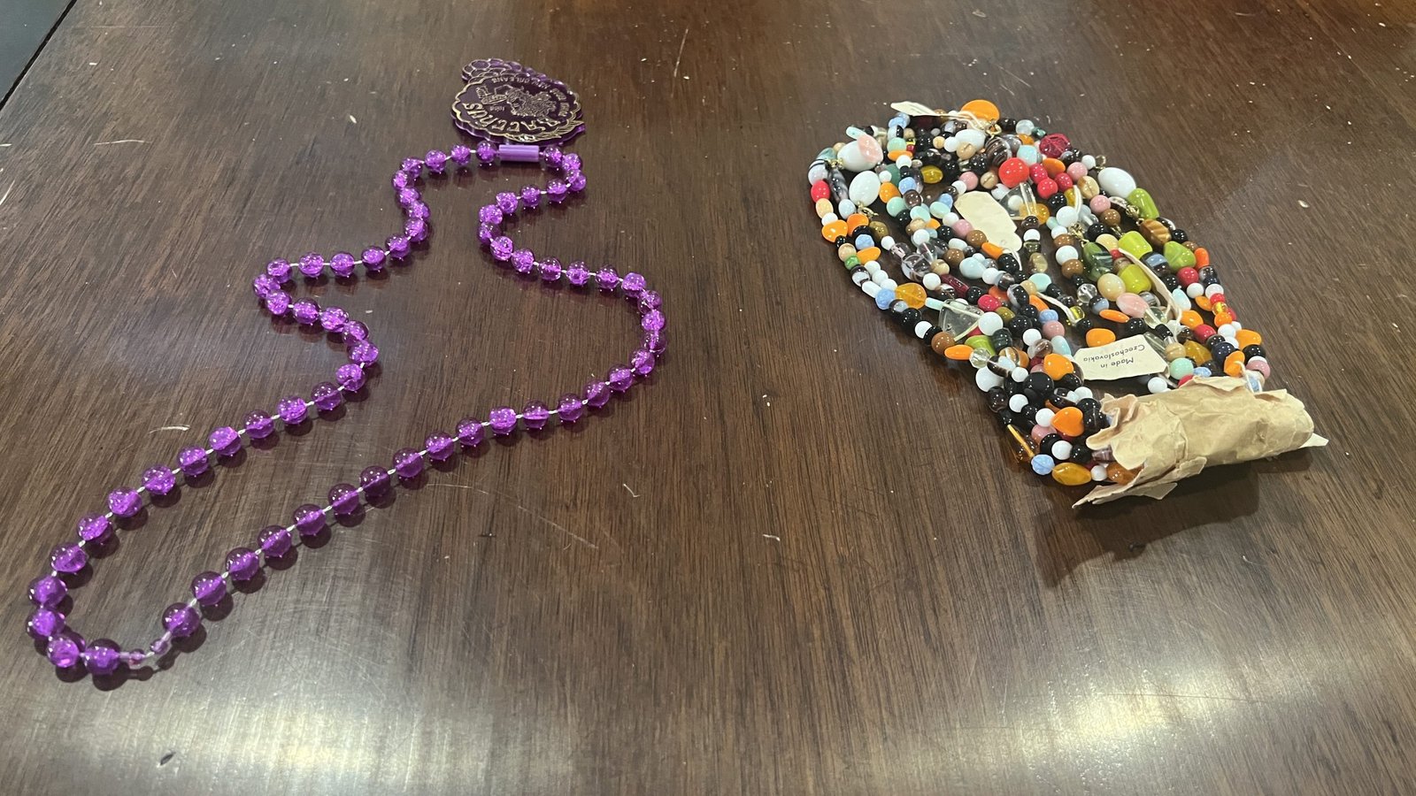 A comparison of two Mardi Gras throws on a wooden surface. On the left, a modern purple beaded necklace with a medallion featuring an embossed design. On the right, a vintage handmade throw, possibly an African-inspired Zulu throw, crafted from multicolored beads strung together in an intricate pattern, with part of the original packaging still attached.