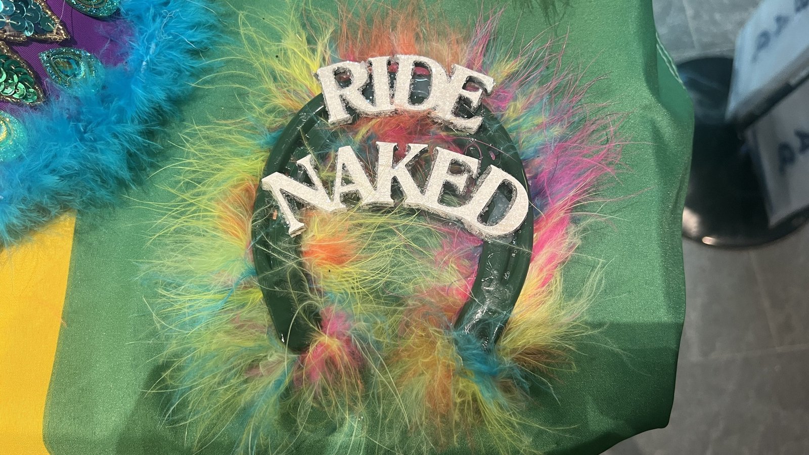 A Mardi Gras horseshoe throw covered in colorful feathers with "RIDE NAKED" in bold white letters, placed on a green tablecloth.
