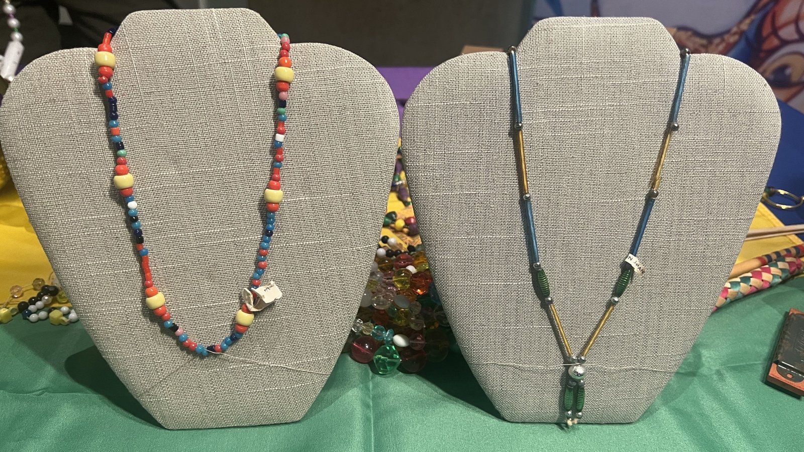 Two handmade Mardi Gras bead necklaces displayed on gray bust stands. The left necklace features multicolored round beads in red, blue, yellow, and white, with a small metallic charm. The right necklace has a more delicate design with blue, gold, and green cylindrical beads, ending in a decorative pendant. Both necklaces are set against a festive Mardi Gras-themed background with scattered beads.
