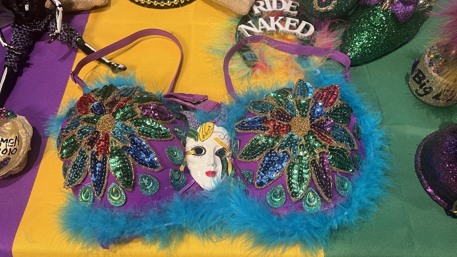 A vibrant Mardi Gras bra throw decorated with peacock-inspired sequins, gold embellishments, and fluffy blue feathers, resting on a purple, green, and gold tablecloth. A small decorative mask is placed on top.