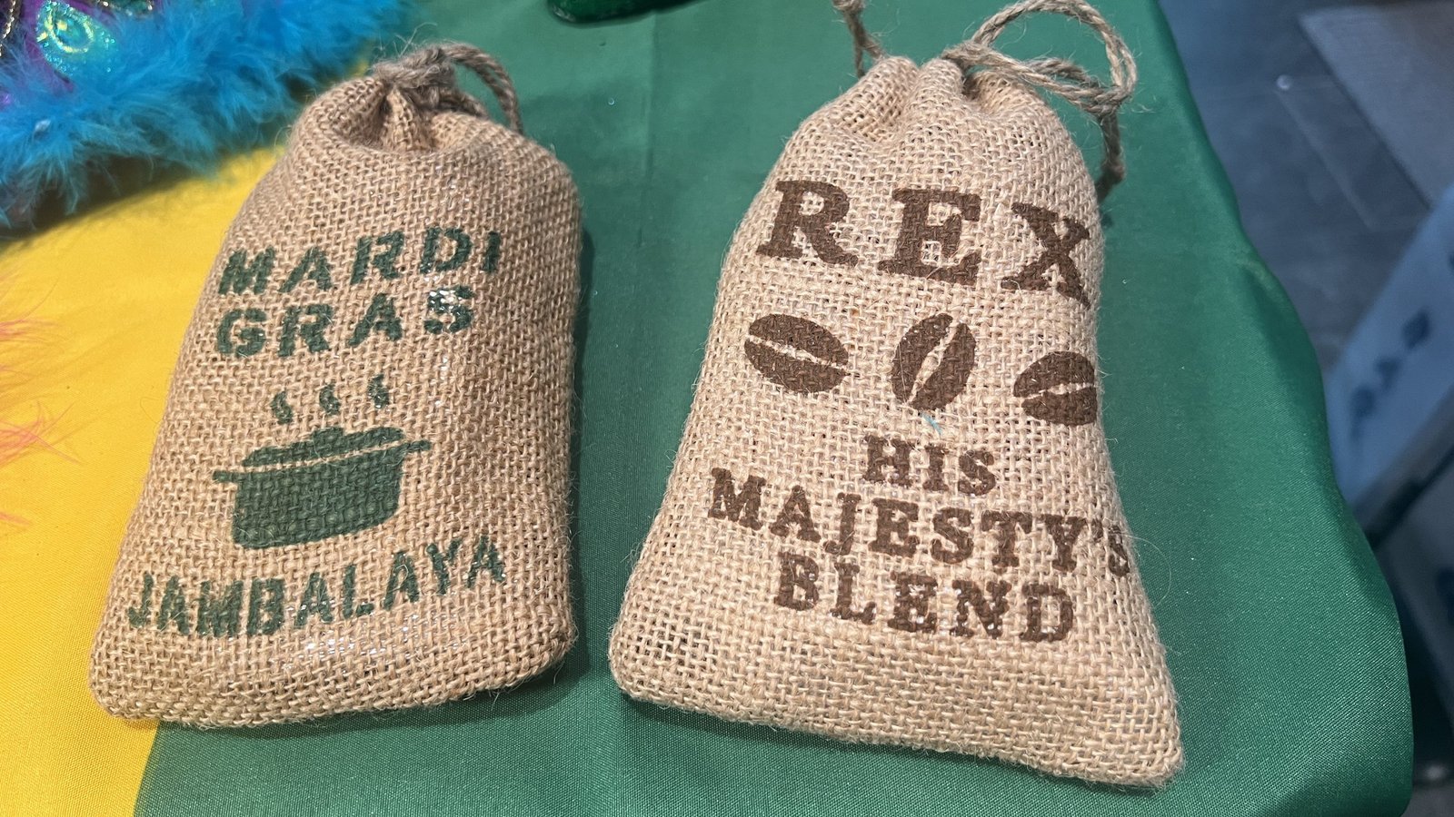 Two burlap sack Mardi Gras throws—one labeled "Mardi Gras Jambalaya" with a pot design, and the other "REX His Majesty’s Blend" with coffee bean graphics—displayed on a green tablecloth.