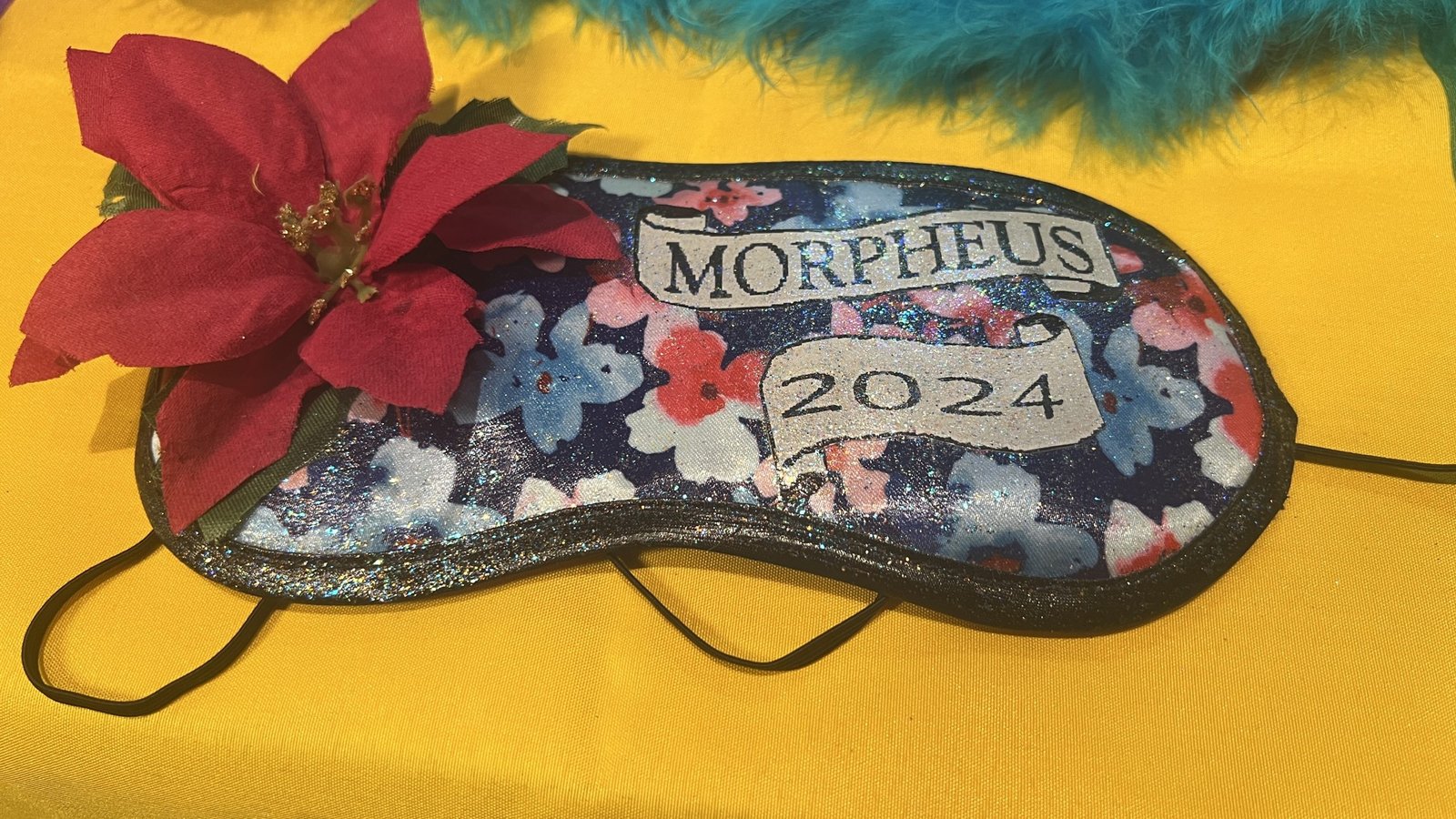 A decorative Mardi Gras sleep mask with "MORPHEUS 2024" written on it, featuring a floral design with blue, pink, and red flowers, plus a red poinsettia embellishment on the side.