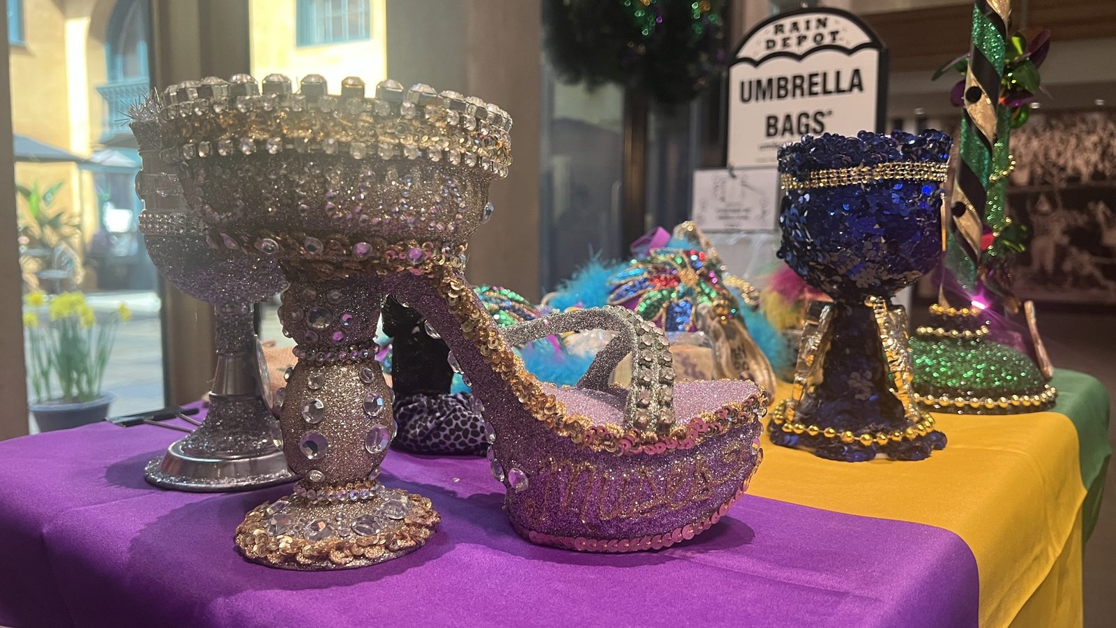 Decorated Mardi Gras throws on a table, including an ornate jeweled goblet and a glittery high-heeled shoe adorned with rhinestones and beads. The background features a festive setting with other decorated items.