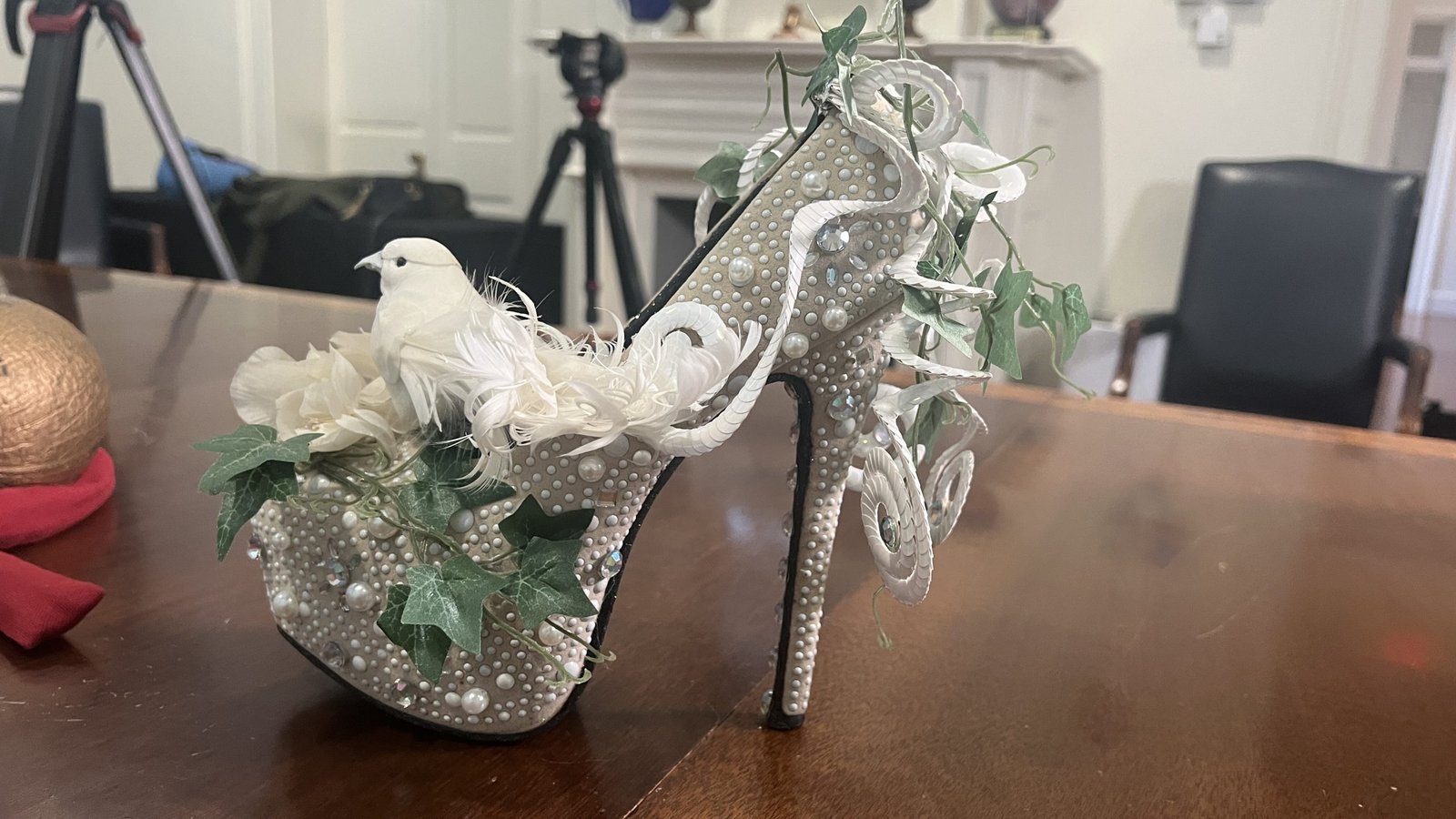 An ornate Mardi Gras shoe throw featuring a high-heeled platform covered in pearls, rhinestones, and decorative ivy. A white dove figurine with feathers sits on the front, adding to the elaborate design. The shoe is displayed on a wooden table, with black chairs and a camera tripod visible in the background.