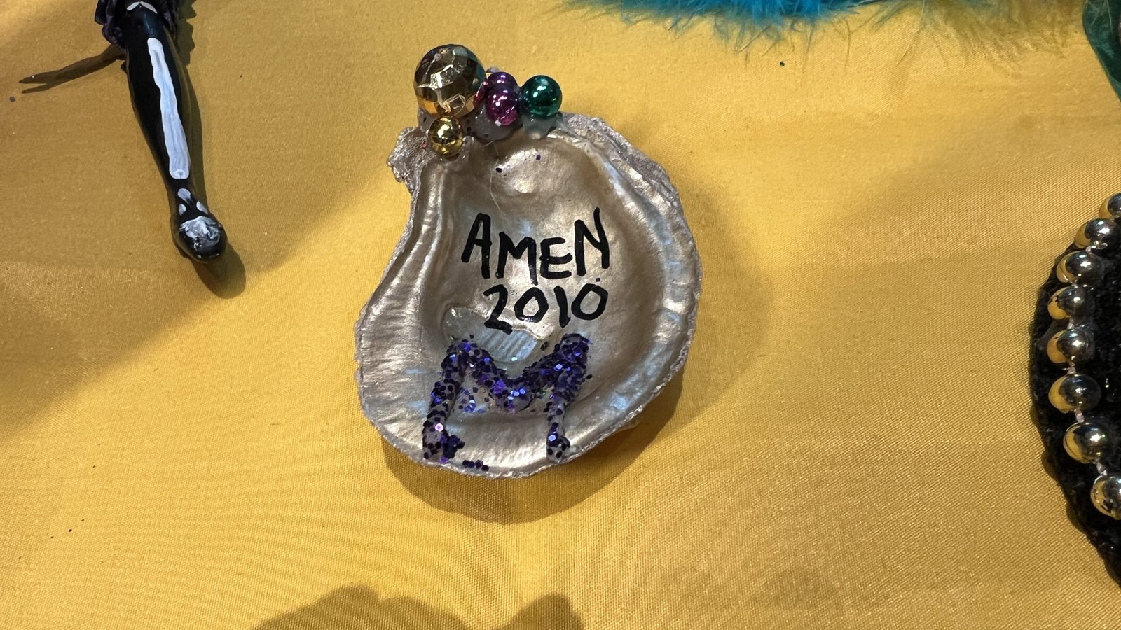 A hand-painted Mardi Gras oyster shell throw with "AMEN 2010" written in black, decorated with purple glitter and small Mardi Gras beads, displayed on a yellow tablecloth.
