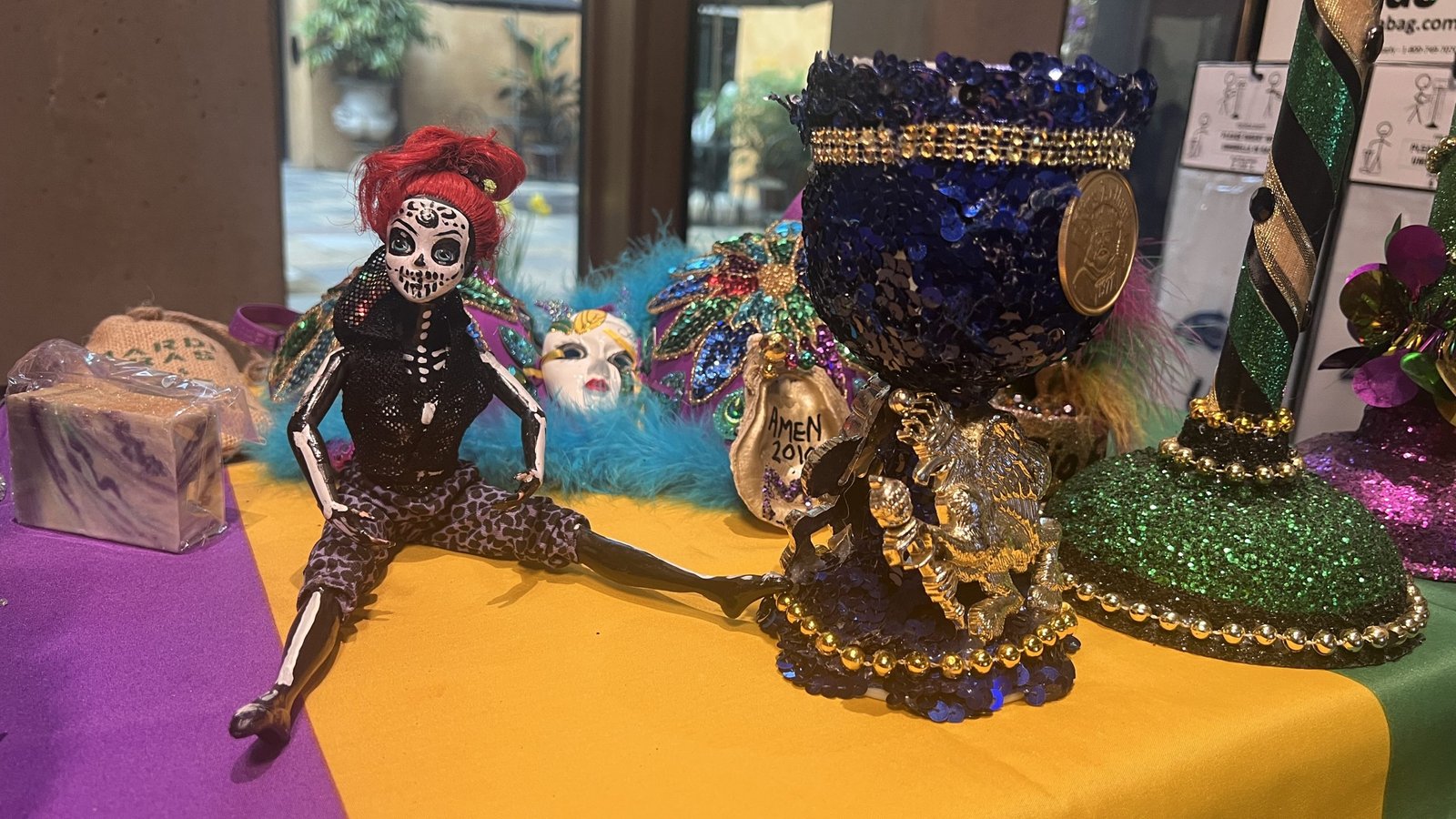 A blue sequined Mardi Gras goblet alongside a skeleton doll with red hair, wearing a black outfit and seated on a festive table. The background includes decorative masks, beads, and other themed items.