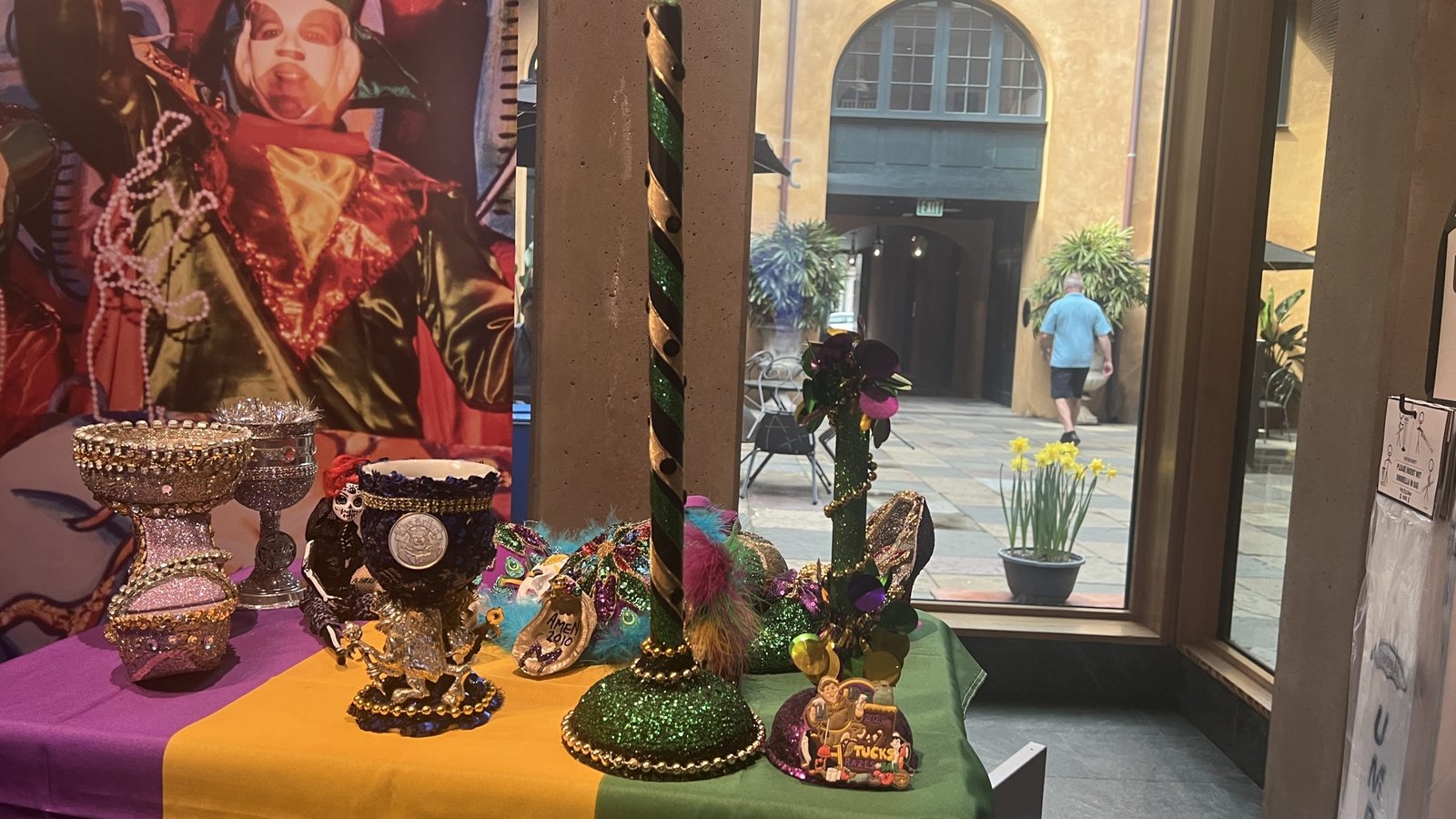 Mardi Gras display with beaded goblets, decorative masks, a glittery green and gold pole, and a small float labeled "THROWS" on a purple, green, and gold tablecloth. A costumed performer’s image is in the background, with a courtyard visible through the window.
