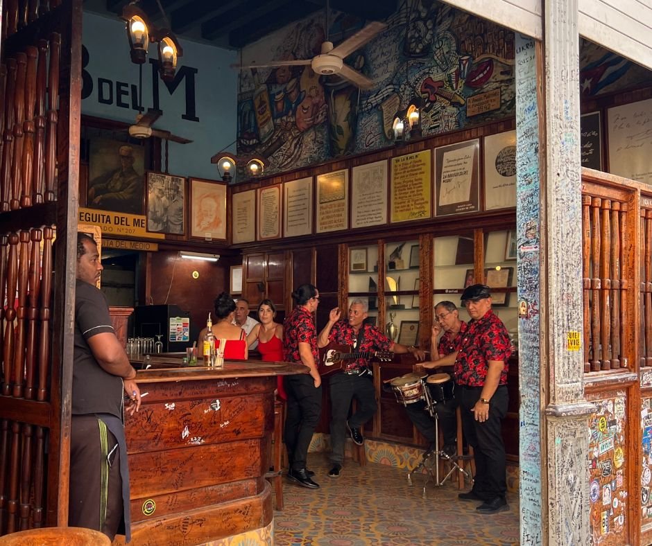 A lively scene inside La Bodeguita del Medio in Havana, Cuba, featuring a traditional Cuban band dressed in red floral shirts performing live music. The rustic wooden bar is covered in handwritten signatures from visitors, while framed photos and writings decorate the walls. A bartender stands behind the bar, while patrons in red dresses enjoy the vibrant atmosphere. The open doorway reveals a graffiti-covered post, adding to the historic and cultural charm of this iconic bar, famous for its mojitos and its connection to Ernest Hemingway.