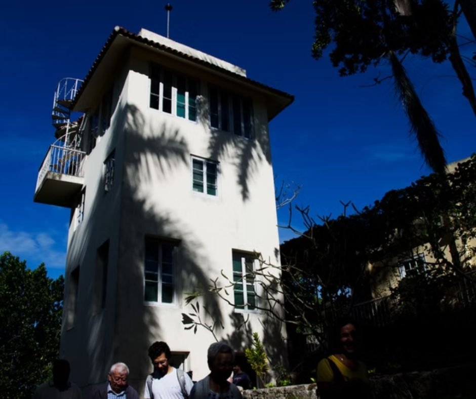 A three-story tower at Finca Vigía, Ernest Hemingway’s former home in Cuba, where he wrote some of his most famous works. The white building stands against a deep blue sky, with a spiral staircase leading to the upper level. Shadows from palm trees add to the atmosphere as visitors explore the grounds.
