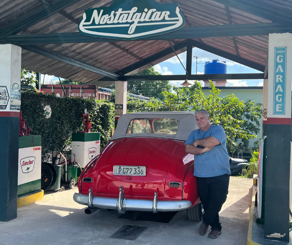 Man leaning against a red vintage convertible at "Nostalgicar" garage, a retro-style gas station in Cuba.