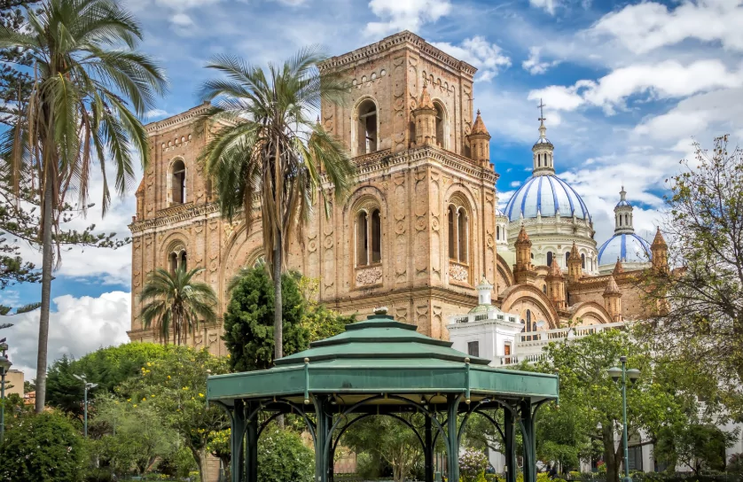 View of the New Cathedral of Cuenca in Ecuador, featuring its ornate brick facade, blue and white domes, and surrounding palm trees and greenery, with a green gazebo in the foreground under a partly cloudy sky.