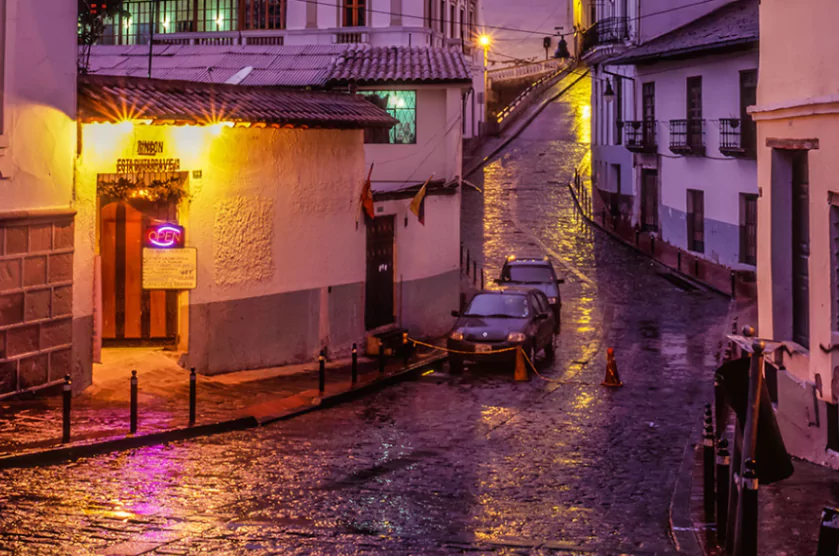 Rain-soaked cobblestone street in Quito’s historic center at dusk, with colonial-style buildings, glowing neon and yellow lights from a corner tavern, and parked cars reflecting on the wet pavement.