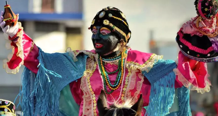 Person in vibrant traditional costume with blackface makeup participating in Ecuador's La Diablada de Píllaro festival; wearing a pink outfit with blue fringe, ornate headwear, and colorful bead necklaces while holding a decorated doll-like figure.