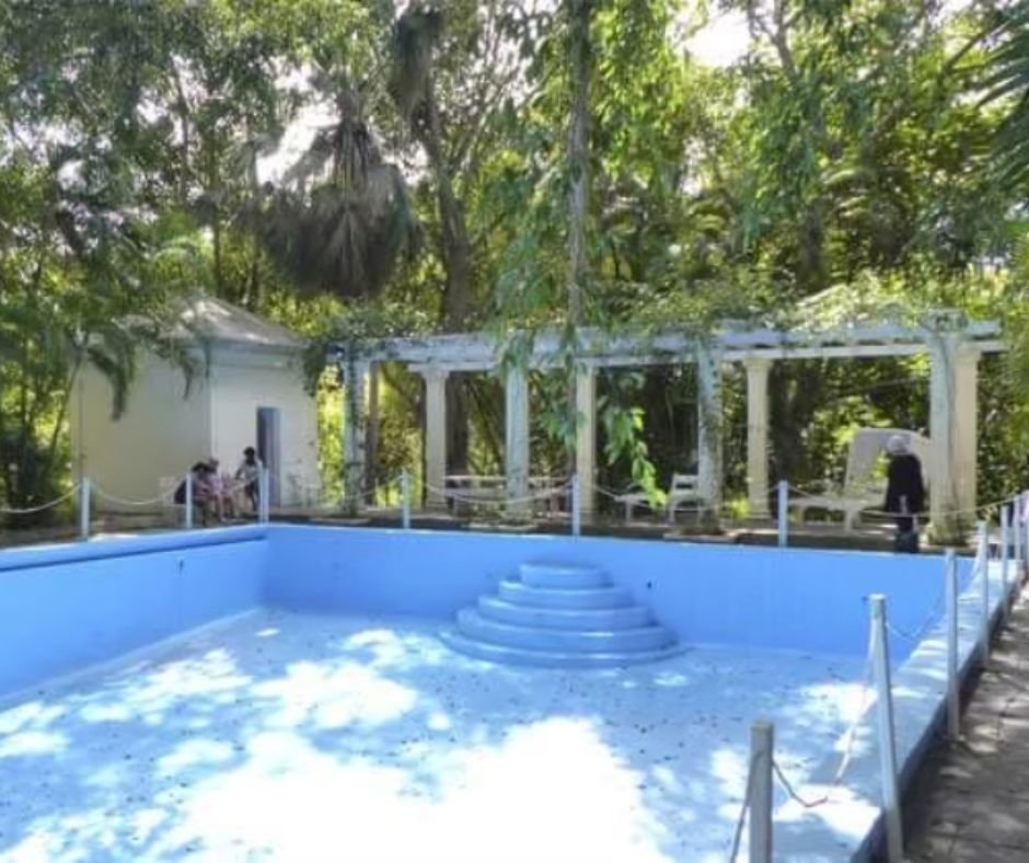 The empty swimming pool at Finca Vigía, once the largest private pool in Cuba, surrounded by lush tropical greenery. A shaded pergola and seating area stand nearby, offering a glimpse into Hemingway’s luxurious yet simple lifestyle.