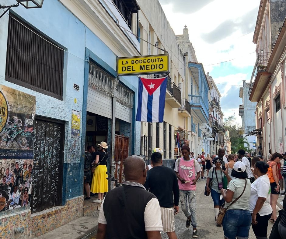 La Bodeguita del Medio exterior – A busy Havana street with people walking past the famous bar, with a Cuban flag hanging outside.