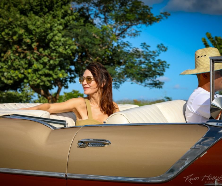 Vintage car ride – A woman in sunglasses enjoying a drive in a classic convertible with a man in a straw hat, evoking mid-century Cuba.