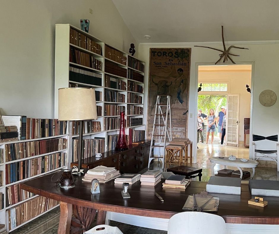 Hemingway’s main library – A literary workspace with bookshelves, a wooden desk, a "Toros en San Sebastián" bullfighting poster, and various artifacts.