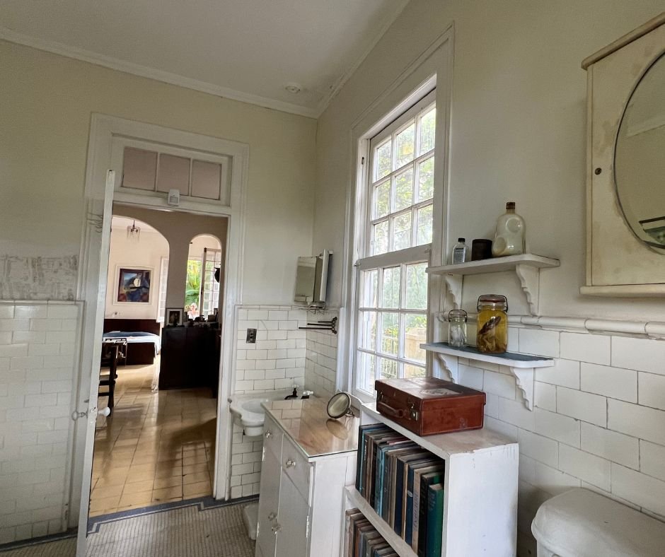 Hemingway’s bathroom – A vintage white-tiled bathroom with bookshelves, glass jars, and a window looking into the house.