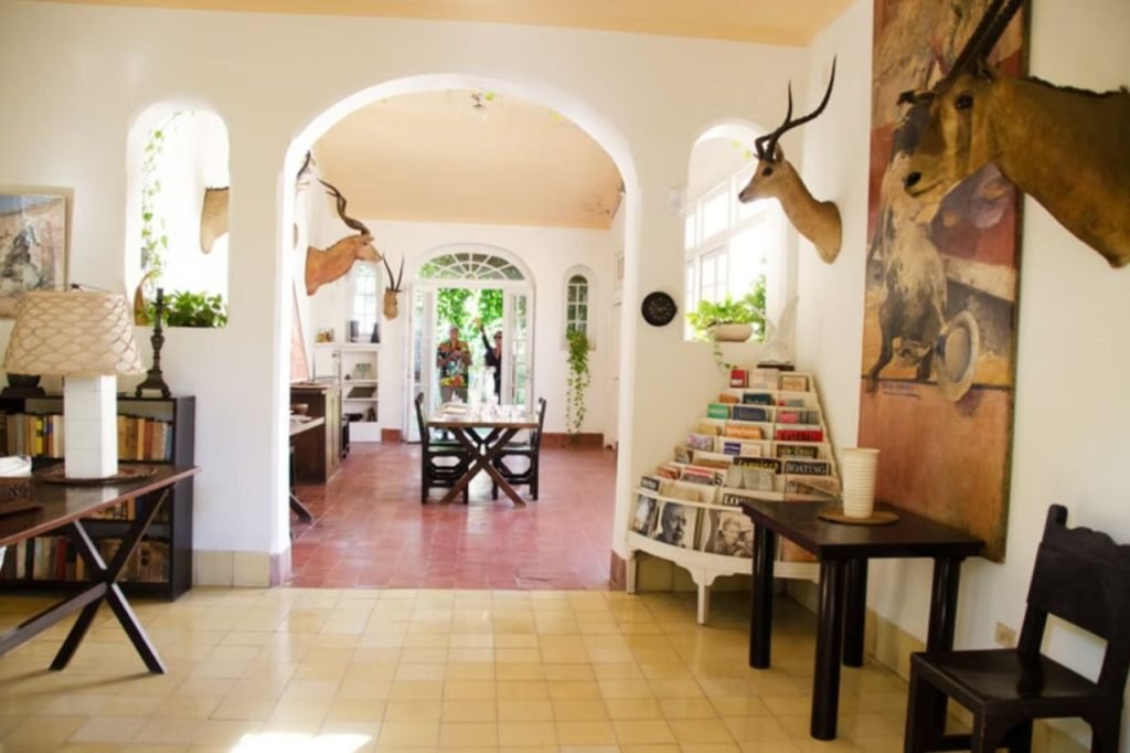 Hemingway's Cuba - A bright and spacious room inside Ernest Hemingway’s Finca Vigía in Cuba, featuring white archways, mounted antelope heads, and a bookshelf filled with magazines and books. The room has a mix of rustic and elegant decor, with wooden furniture, tiled floors, and natural light streaming in through large windows. In the background, visitors can be seen exploring the home, which remains preserved as a museum showcasing Hemingway’s life and legacy.