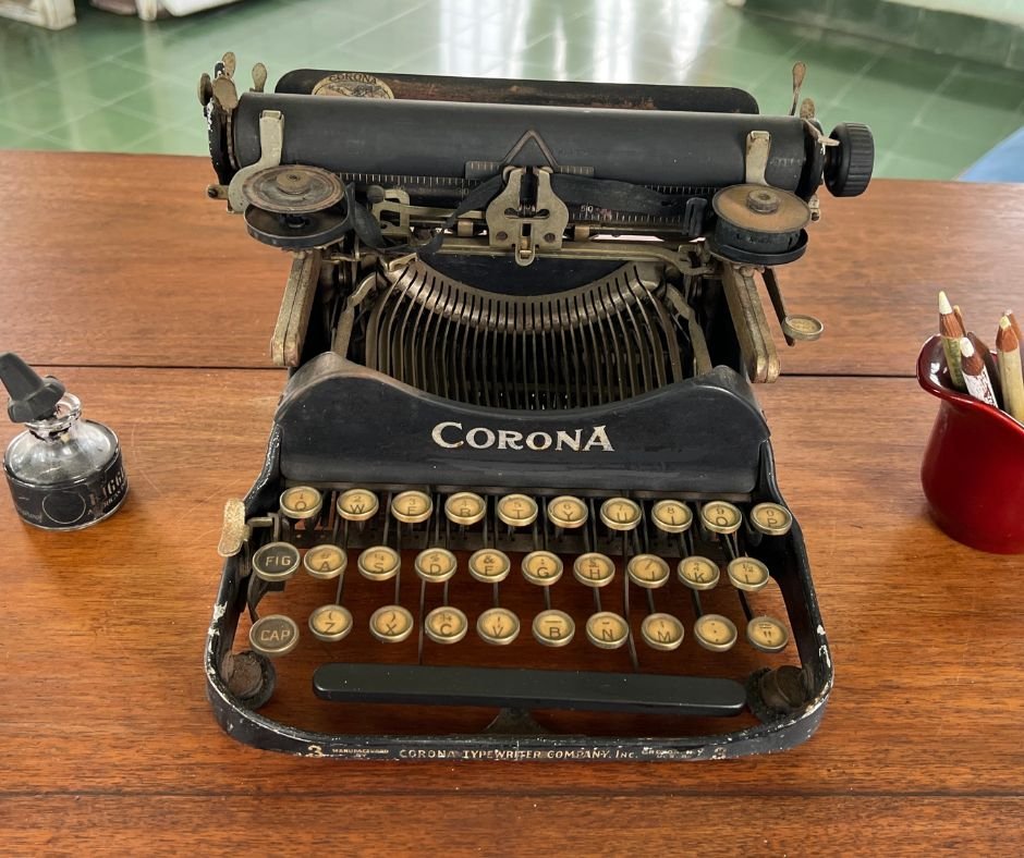 Hemingway’s typewriter – A vintage black Corona typewriter on a wooden desk, accompanied by an ink bottle and a red container with pencils, reflecting the writer’s workspace.