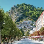 A picturesque view of Berat, Albania, showcasing its traditional white Ottoman-style houses stacked on a hillside beneath a fortress perched on a rocky peak. The foreground features a pedestrian-friendly street lined with lush green trees, street lamps, and outdoor cafes, with a clear blue sky above.