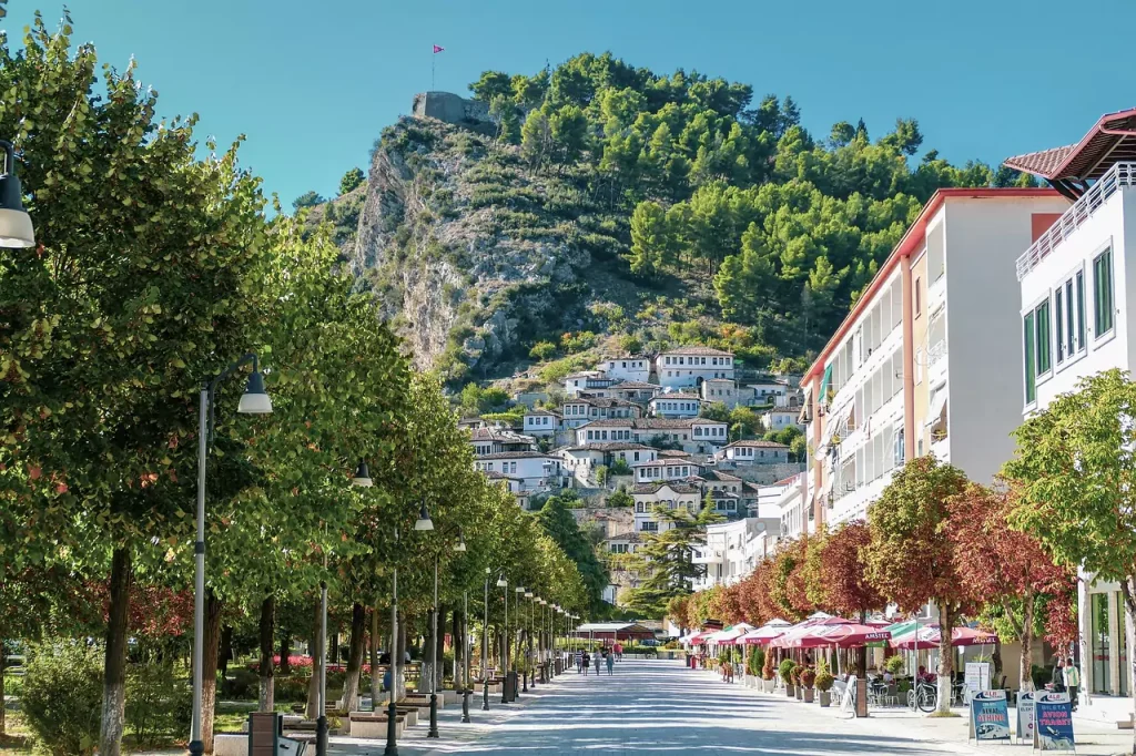 A picturesque view of Berat, Albania, showcasing its traditional white Ottoman-style houses stacked on a hillside beneath a fortress perched on a rocky peak. The foreground features a pedestrian-friendly street lined with lush green trees, street lamps, and outdoor cafes, with a clear blue sky above.