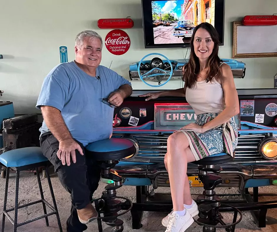 Man and woman sitting on bar stools made from car parts in an automotive-themed garage, with a vintage Chevrolet front grille as décor and classic car memorabilia on the walls. A TV screen in the background displays an image of classic cars in Cuba.