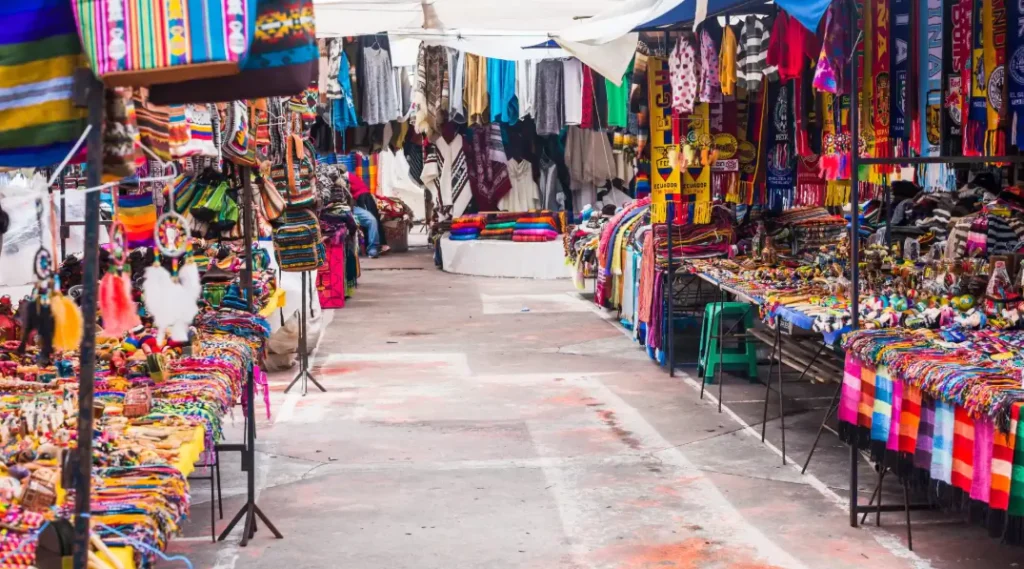 Colorful outdoor market in Otavalo, Ecuador, featuring rows of vibrant textiles, handmade jewelry, traditional scarves, ponchos, bags, and souvenirs displayed under canopies along a narrow walkway.